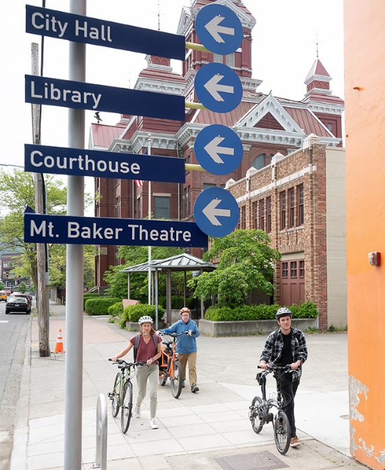 Two people walking their bicycles near a street sign in downtown Bellingham, pointing to City Hall, Library, Courthouse, and Mt. Baker Theatre.